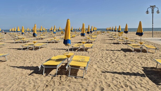A beach with rows of yellow and blue beach chairs and umbrellas, a clear sky, and a calm sea.