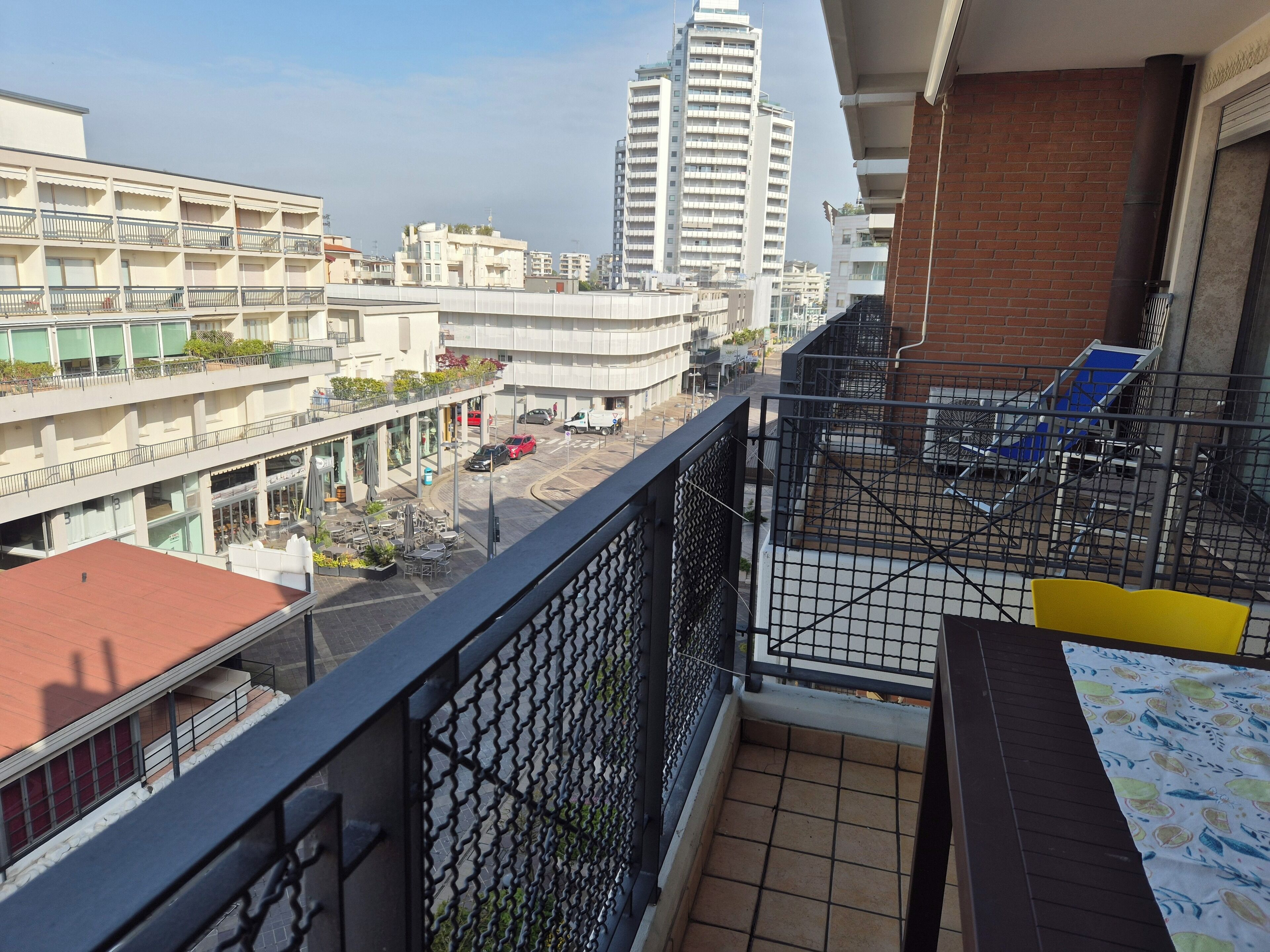 Un balcón con vistas a un paisaje urbano, que incluye un edificio alto, una zona comercial y una silla azul.