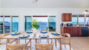 A dining area with a view of the ocean, wooden furniture, and a kitchen with wooden cabinets. - Paradise Ocean Vista- Absolute Oceanfront gorgeous views, A/C & laundry! (Keaau)