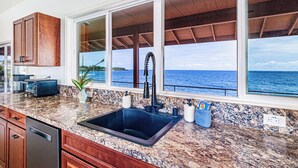 A kitchen with granite countertops, a black sink, wooden cabinets, and large windows overlooking the ocean. - Paradise Ocean Vista- Absolute Oceanfront gorgeous views, A/C & laundry! (Keaau)