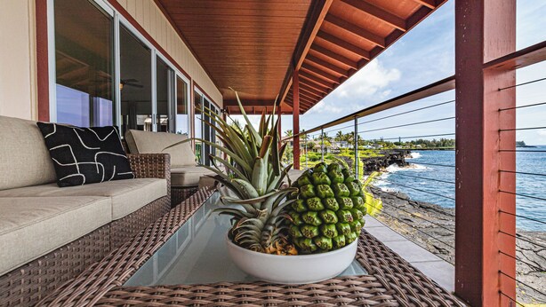 A covered outdoor seating area with a wicker table, a bowl of pineapple, and a sofa with a black pillow. - Paradise Ocean Vista- Absolute Oceanfront gorgeous views, A/C & laundry! (Keaau)