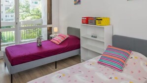 A bedroom with two beds, a window with a view of buildings and trees, a white shelf with colorful items, and a 'HAPPY' framed picture on the wall. - Charming T4 with parking and terrace - sleeps up to 6 (Avignon)