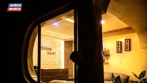 A view through a window of a hotel room with a visible bed, a wooden headboard, and a wall with framed pictures. - Mirton Hotel (Bujumbura)