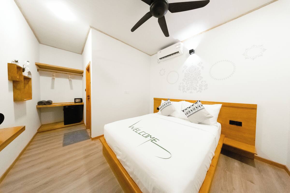 A hotel room with a bed, a ceiling fan, an air conditioning unit, a wooden headboard, and a welcome sign on the bed.