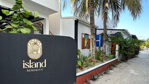 En skylt med logotypen och namnet ”Island Serenity” framför en byggnad med en vit fasad, palmer och en röd blomlåda längs trottoaren.