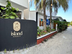 A sign with the logo and name "Island Serenity" in front of a building with a white facade, palm trees, and a red planter along the sidewalk. - Island Serenity  (Dhiffushi)