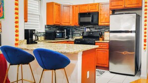 Una cocina con armarios naranjas, un refrigerador de acero inoxidable, un microondas y una cafetera. Hay dos taburetes azules y un salpicadero de azulejos.