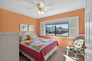 Un dormitorio con cama, ventana con contraventanas, ventilador de techo y vista a casas exteriores.
