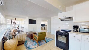 Una cocina moderna con estufa, horno y cafetera, un comedor con mesa y sillas, y una sala de estar con sofá y plantas decorativas.