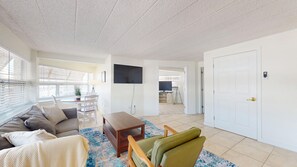 A living room with a sofa, a coffee table, and a TV. There is a door and a window with blinds.