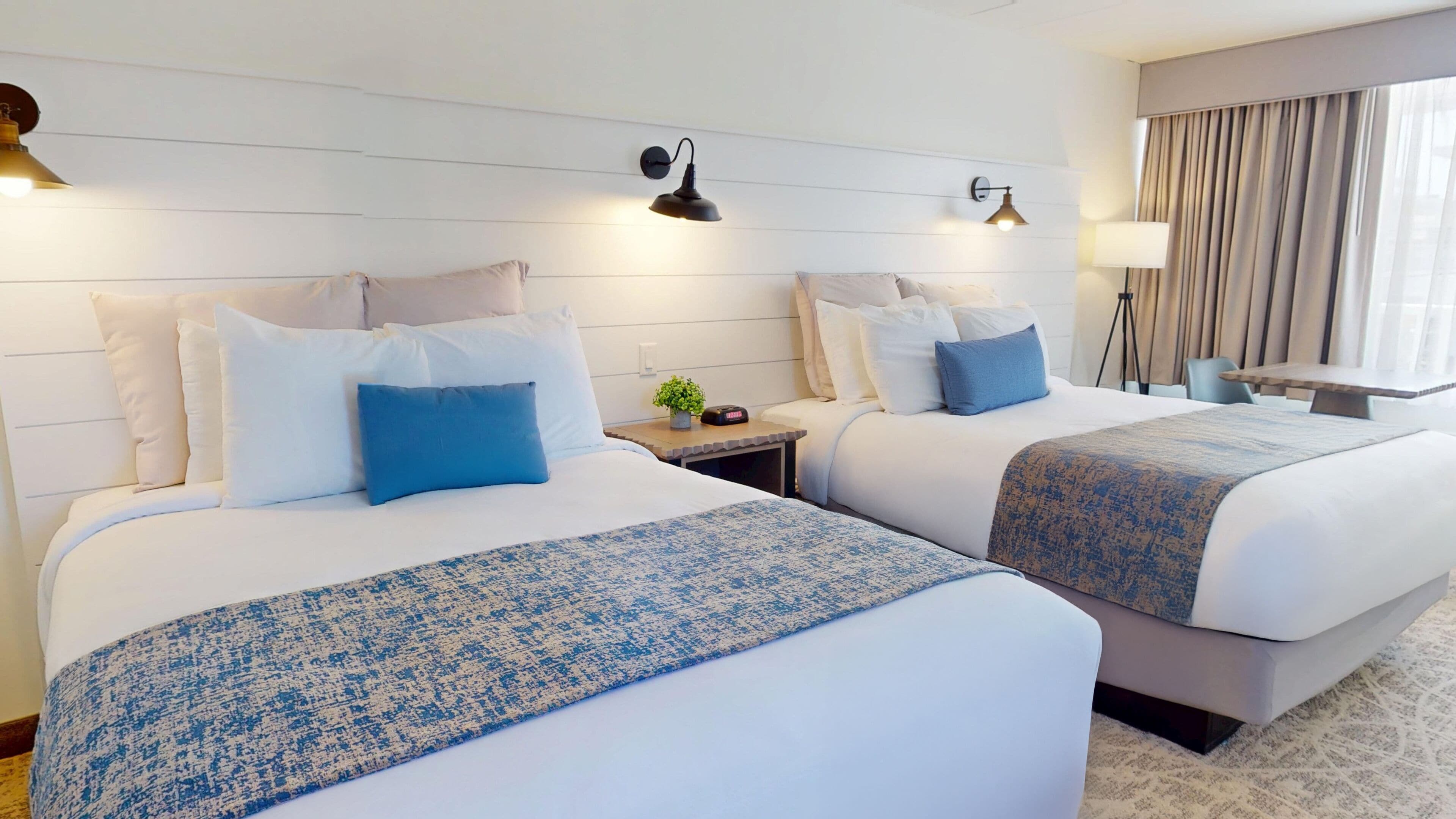 Quarto de hotel com duas camas, uma mesa de cabeceira com relógio, uma luminária e vista para o exterior pela janela.