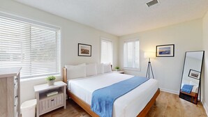 Quarto com cama, mesas de cabeceira, abajur, espelho e janelas com persianas.
