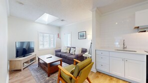 A modern living room with a flat-screen TV, a sofa, a coffee table, and a kitchen area with white cabinets and a sink. - King Suite Upper (502) (Ocean City)