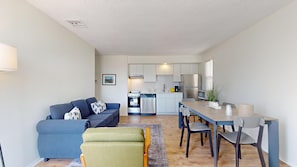 Ein modernes Wohnzimmer mit einem blauen Sofa, einem grünen Sessel und einem Esstisch mit Stühlen. Im Hintergrund ist eine Küche mit weißen Schränken und Geräten zu sehen.