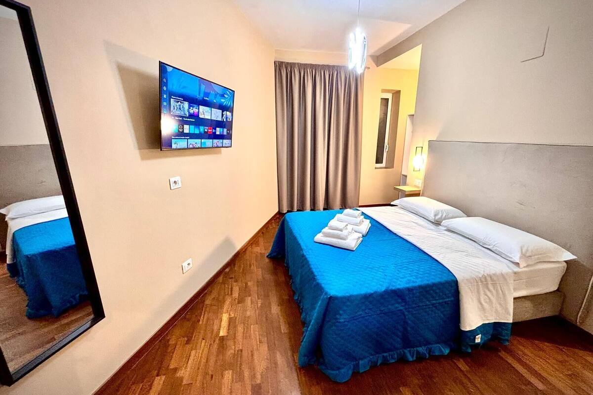 Habitación de hotel con cama, televisor montado en la pared, espejo y ventana con cortinas.