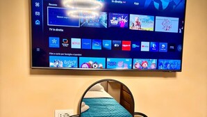 Ein Hotelzimmer mit Fernseher, der Streaming-Dienste anzeigt, einem Nachttisch mit Kaffeemaschine, Teebeuteln, einer Taschentuchbox und einer Champagnerflasche.