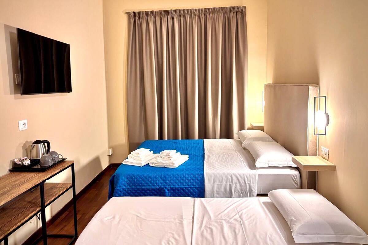 Una habitación de hotel con cama, dos mesitas de noche, un escritorio con hervidor y vasos, televisor y un ventanal con cortinas.