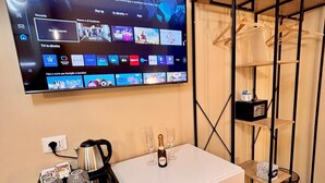 Ein Hotelzimmer mit einem an der Wand befestigten Flachbildfernseher, der verschiedene Streaming-Dienste anzeigt. Es gibt eine weiße Minibar mit einer Kaffeemaschine, Gläsern und einer Champagnerflasche. Außerdem ist ein Metallregal mit einer Lampe und einer kleinen Schachtel zu sehen.