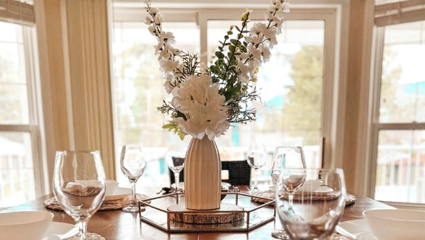 Una mesa de comedor dispuesta con platos, cuencos y cubiertos blancos, con un jarrón de flores blancas en el centro.