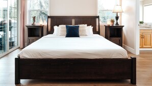Una cama grande con estructura de madera oscura, ropa de cama blanca y varios almohadones. Hay dos mesitas de noche con lámparas y objetos decorativos. La habitación tiene ventanas amplias con vistas a árboles y una terraza afuera.