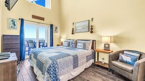 A bedroom with a large bed, a desk, a chair, a nightstand, and a view of the ocean.
