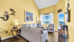 A living room with a sofa, armchairs, a small table with a lamp, and a view of the ocean through large windows.
