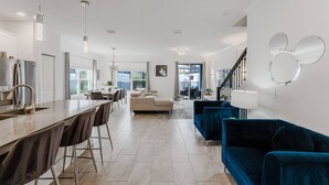 A modern kitchen with a large island, a dining area with chairs, a living room with a blue sofa, and a staircase leading to another level.