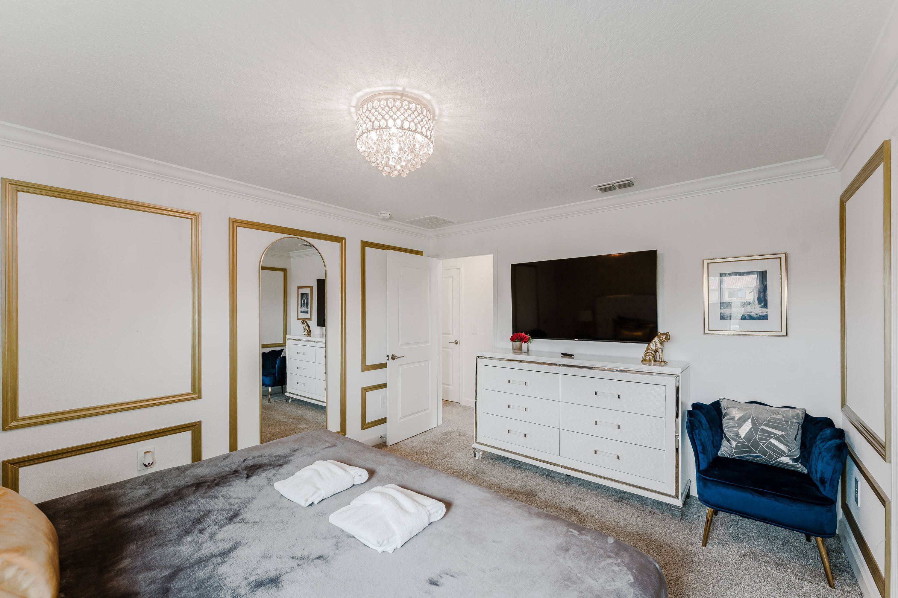 Une chambre avec un grand lit, une commode, une coiffeuse, un téléviseur et un fauteuil bleu.