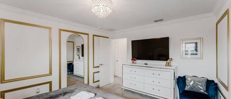 Une chambre avec un grand lit, une commode, une coiffeuse, un téléviseur et un fauteuil bleu.