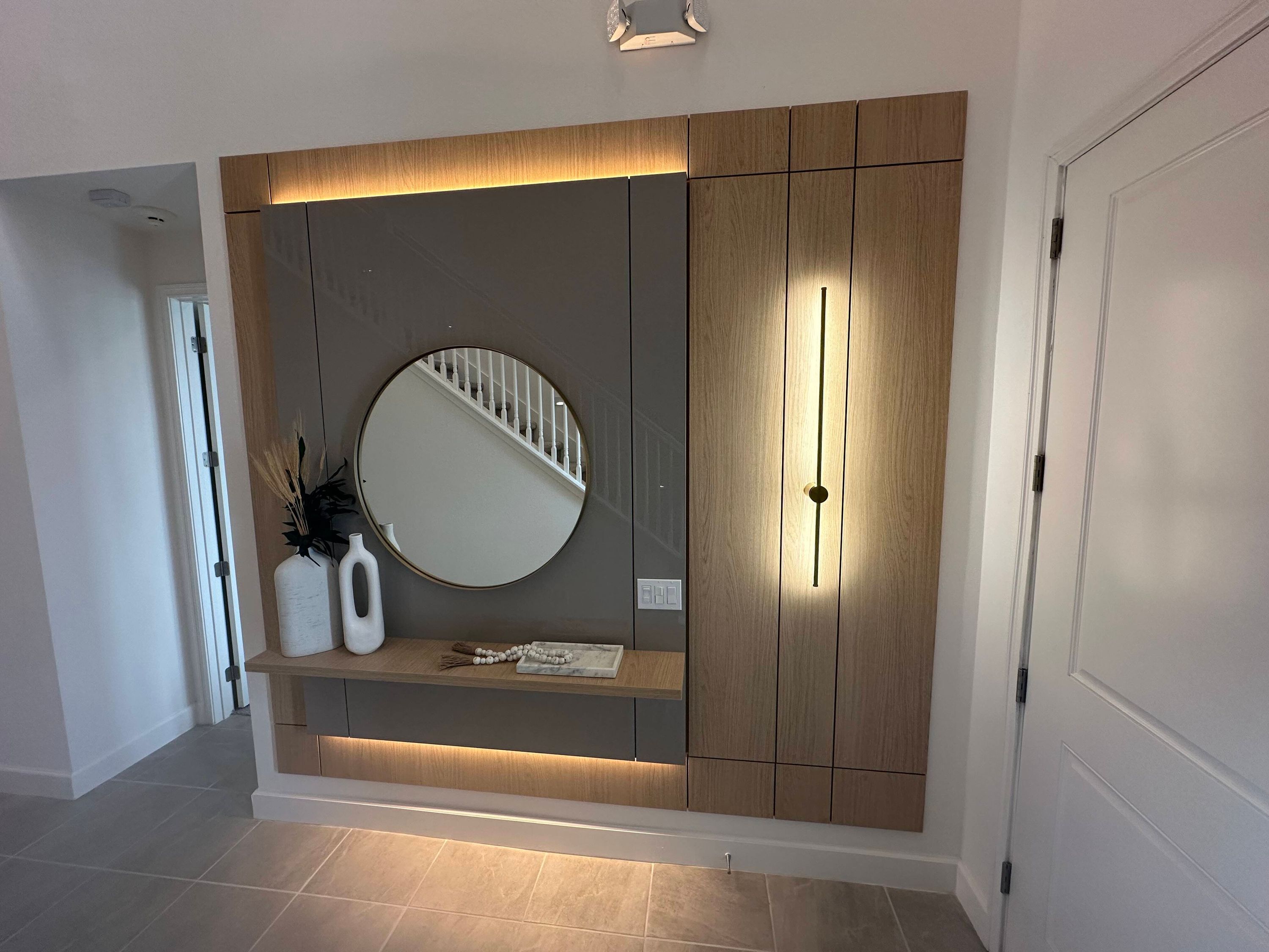 A modern interior with a built-in wooden cabinet, a round mirror, and a shelf with decorative items.