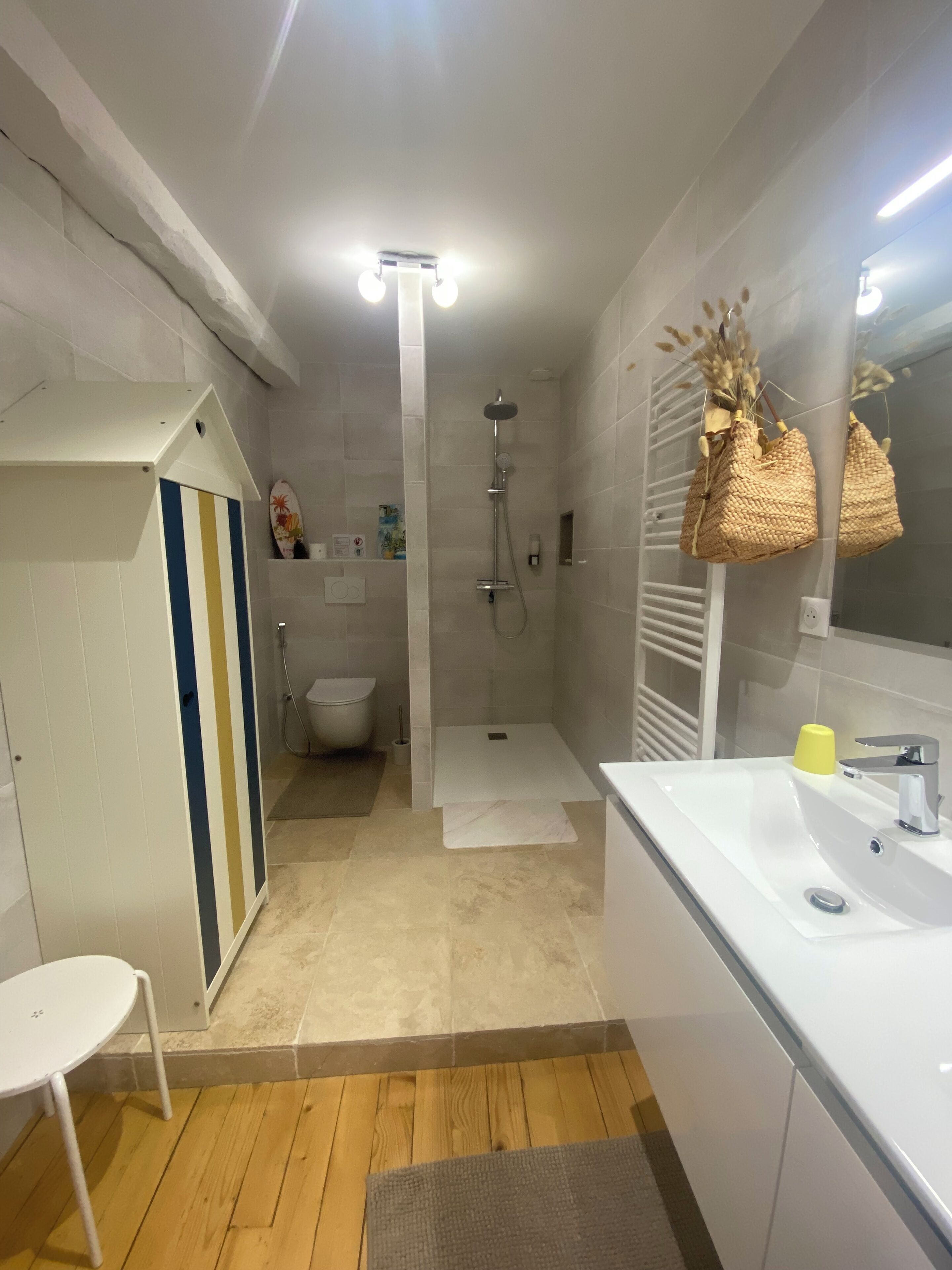 Une salle de bain moderne avec une douche, des toilettes et un lavabo. Le sol est en bois, il y a une chaise blanche et un porte-serviettes.