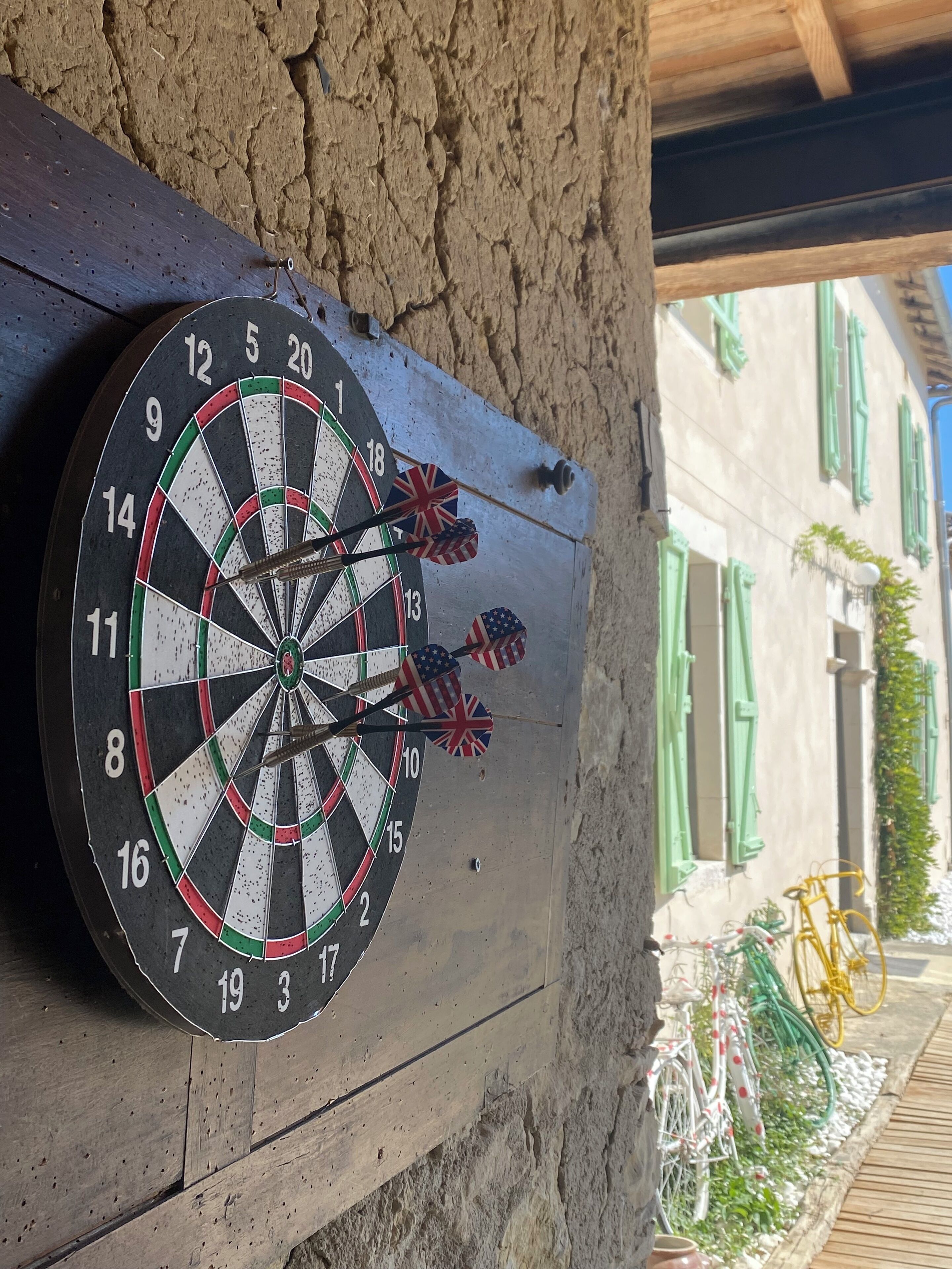 Un jeu de fléchettes fixé au mur, qui présente une texture rustique, un vélo appuyé contre le mur et, au fond, un bâtiment avec des volets verts.
