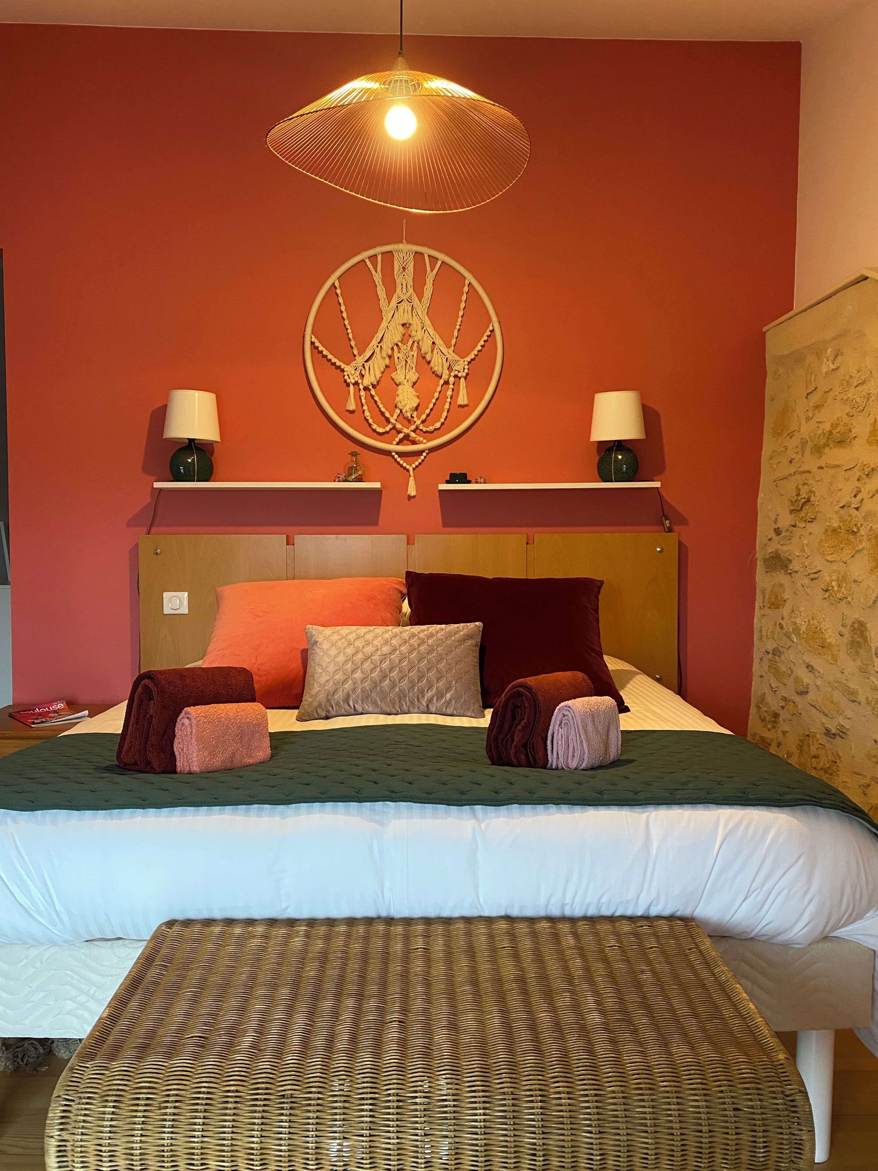 A double bed with a green bedspread, two pillows, and a headboard. There is a wall hanging above the bed and a lamp on a shelf.
