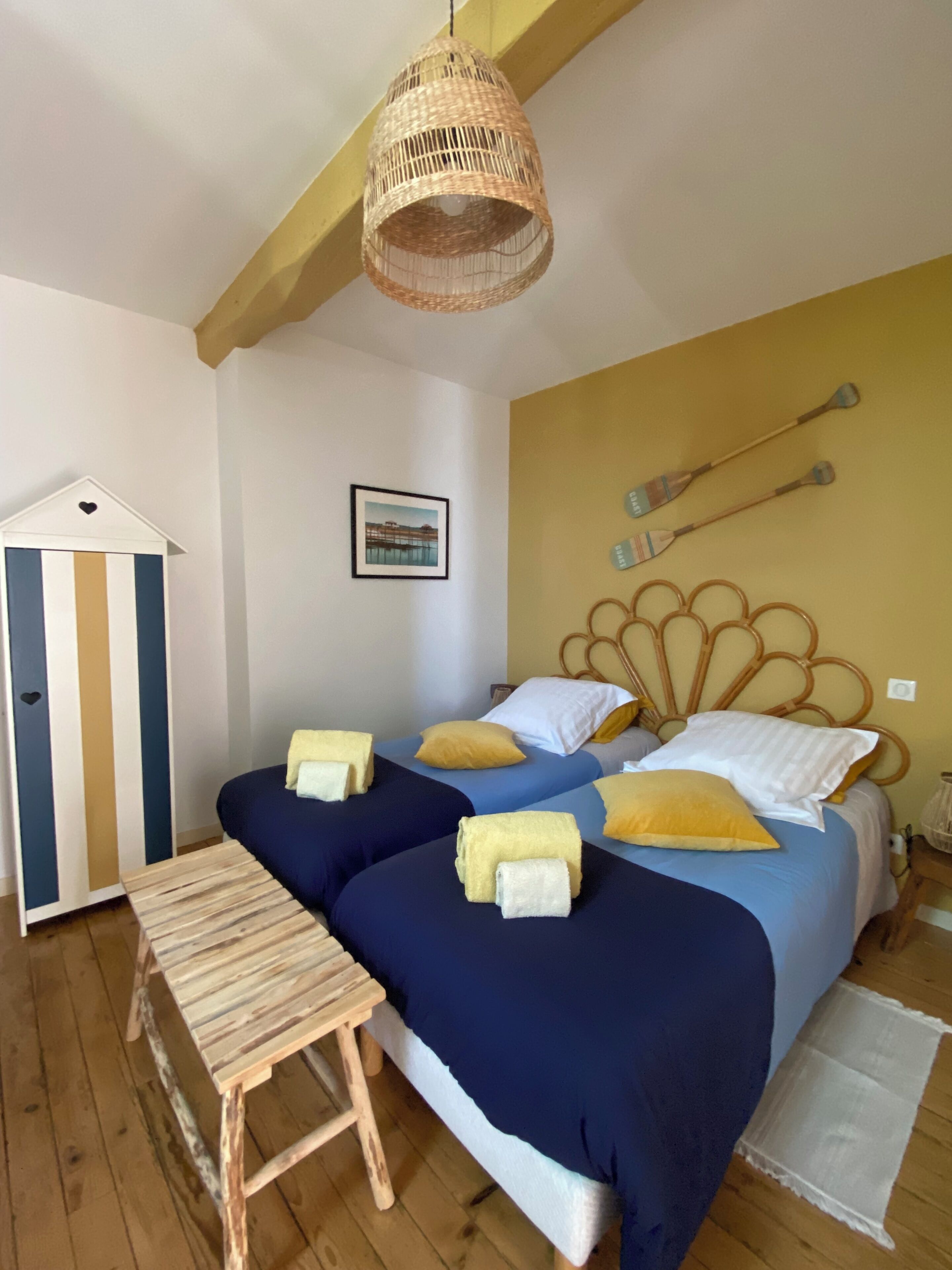 Un dormitorio con dos camas, un banco de madera, una lámpara colgante de mimbre, una pared a rayas y un cuadro enmarcado.