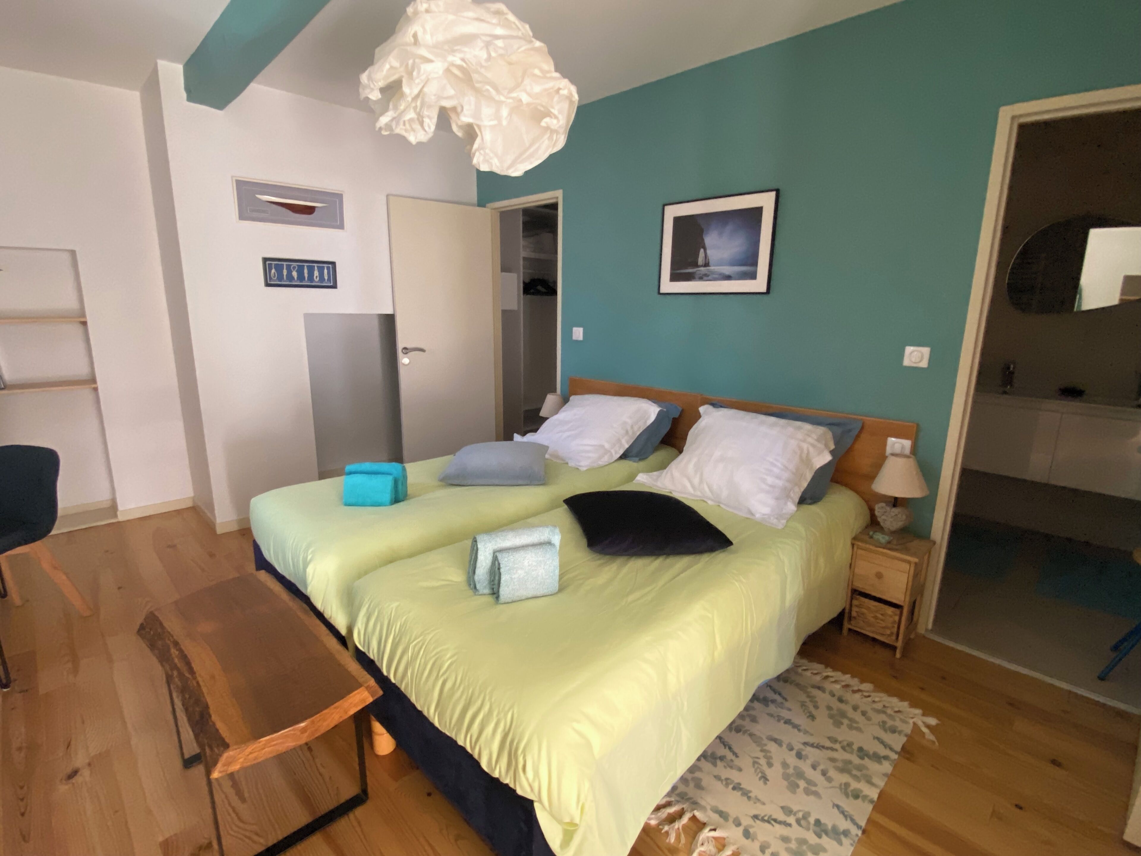 A bedroom with a bed, bedside tables, a wooden bench, a hanging light fixture, a wall-mounted picture, and a door leading to a bathroom.