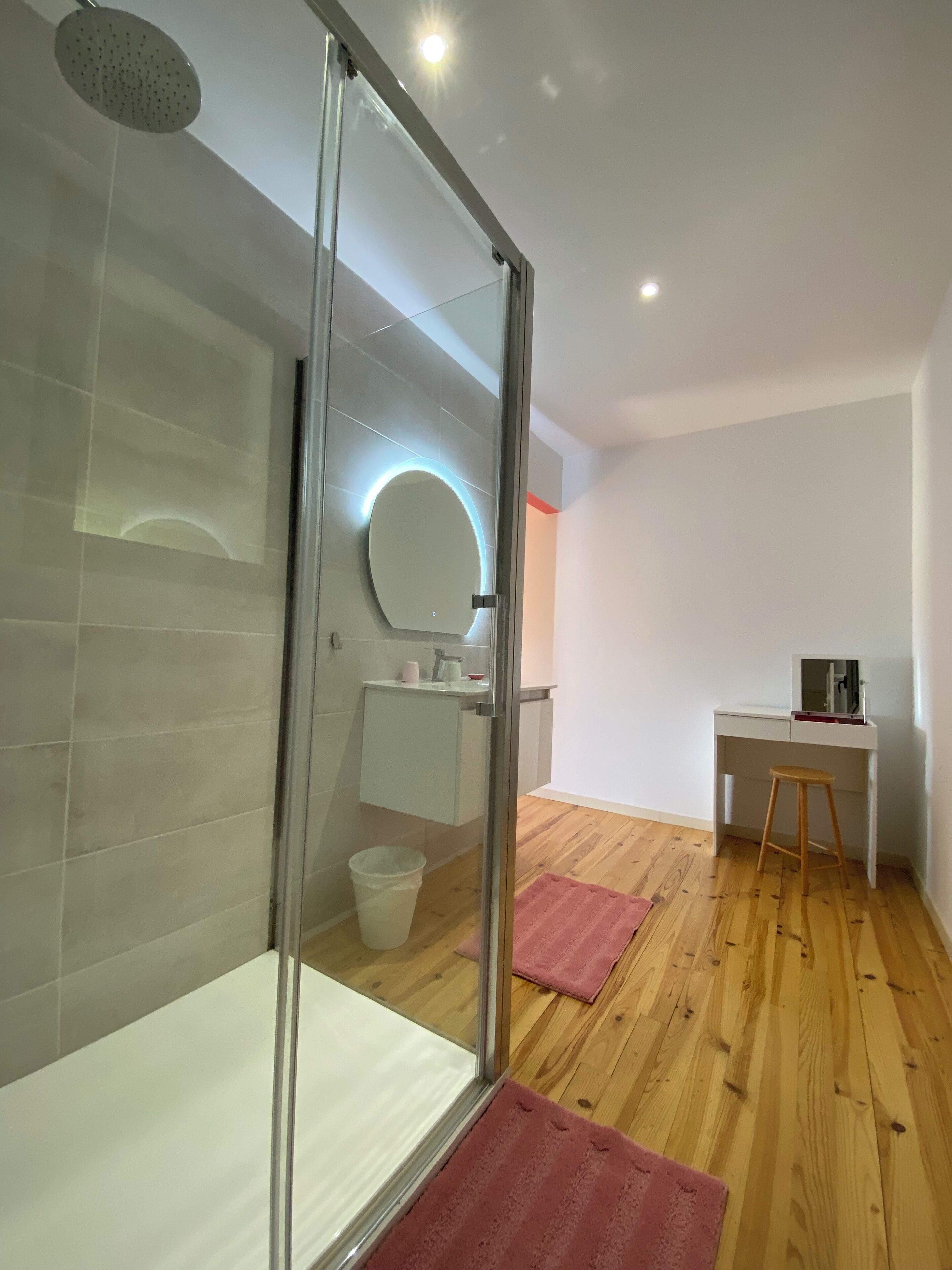 A modern bathroom with a glass shower enclosure, a round mirror with a light, a white sink, and a wooden floor.