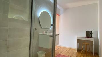 A modern bathroom with a glass shower enclosure, a round mirror with a light, a white sink, and a wooden floor.