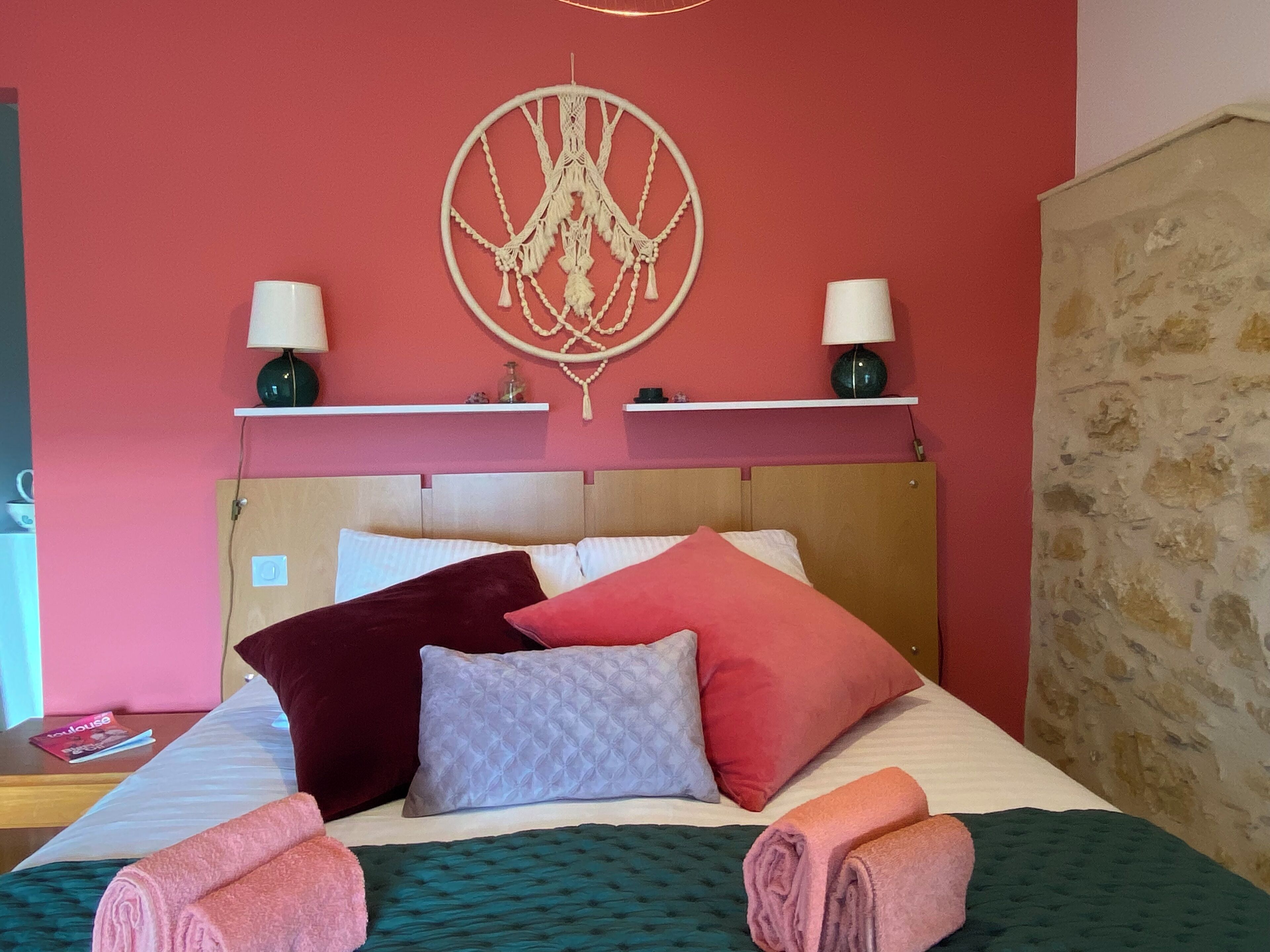 Una cama bien tendida con varias almohadas y toallas enrolladas, un adorno de macramé en la pared, y dos lámparas de noche sobre una estantería.