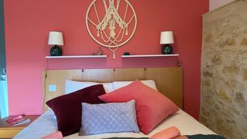 Una cama bien tendida con varias almohadas y toallas enrolladas, un adorno de macramé en la pared, y dos lámparas de noche sobre una estantería.