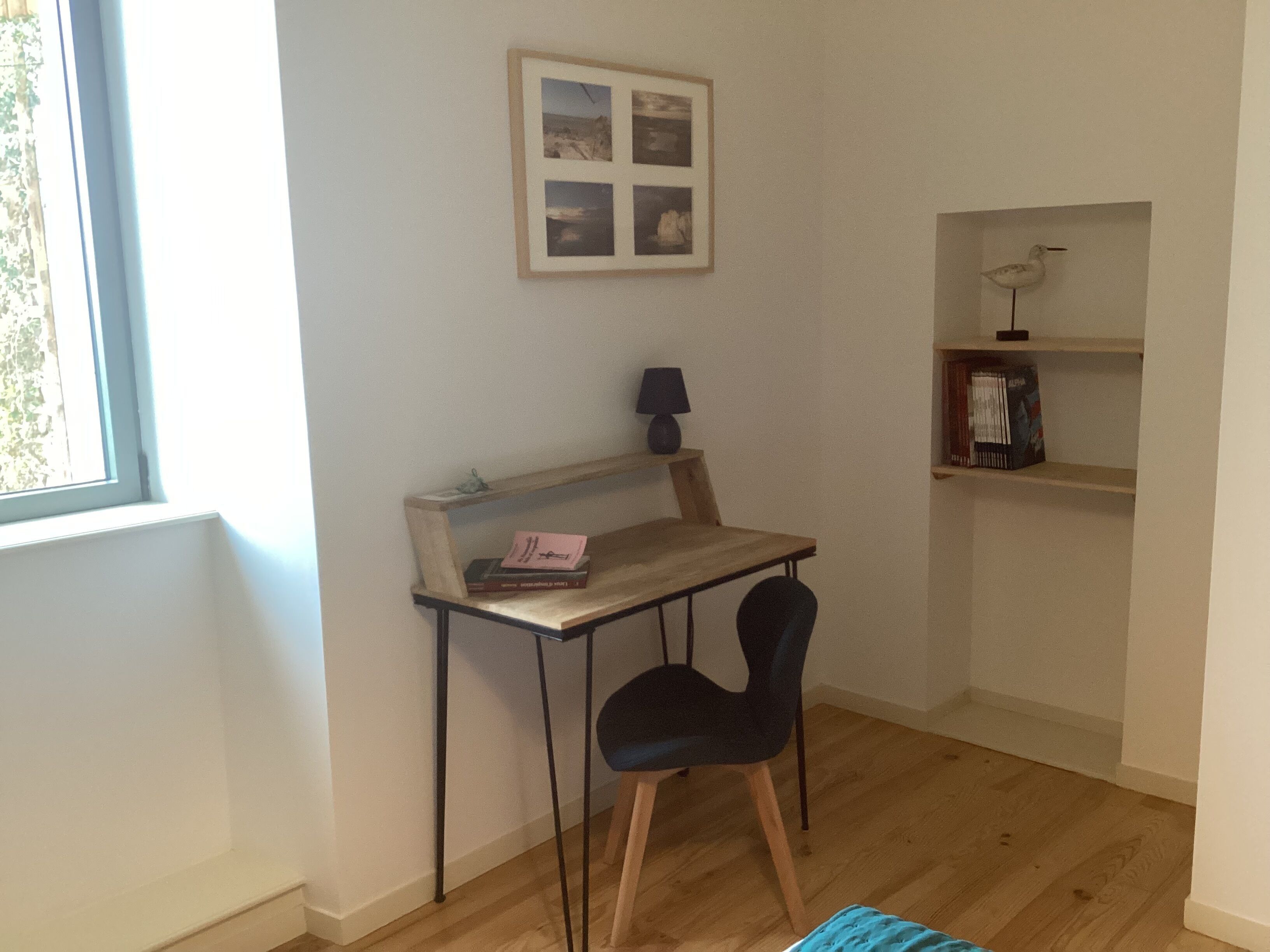 Una habitación pequeña y bien iluminada, con un escritorio de madera, una silla negra, un cuadro en la pared y una estantería con libros y una lámpara.