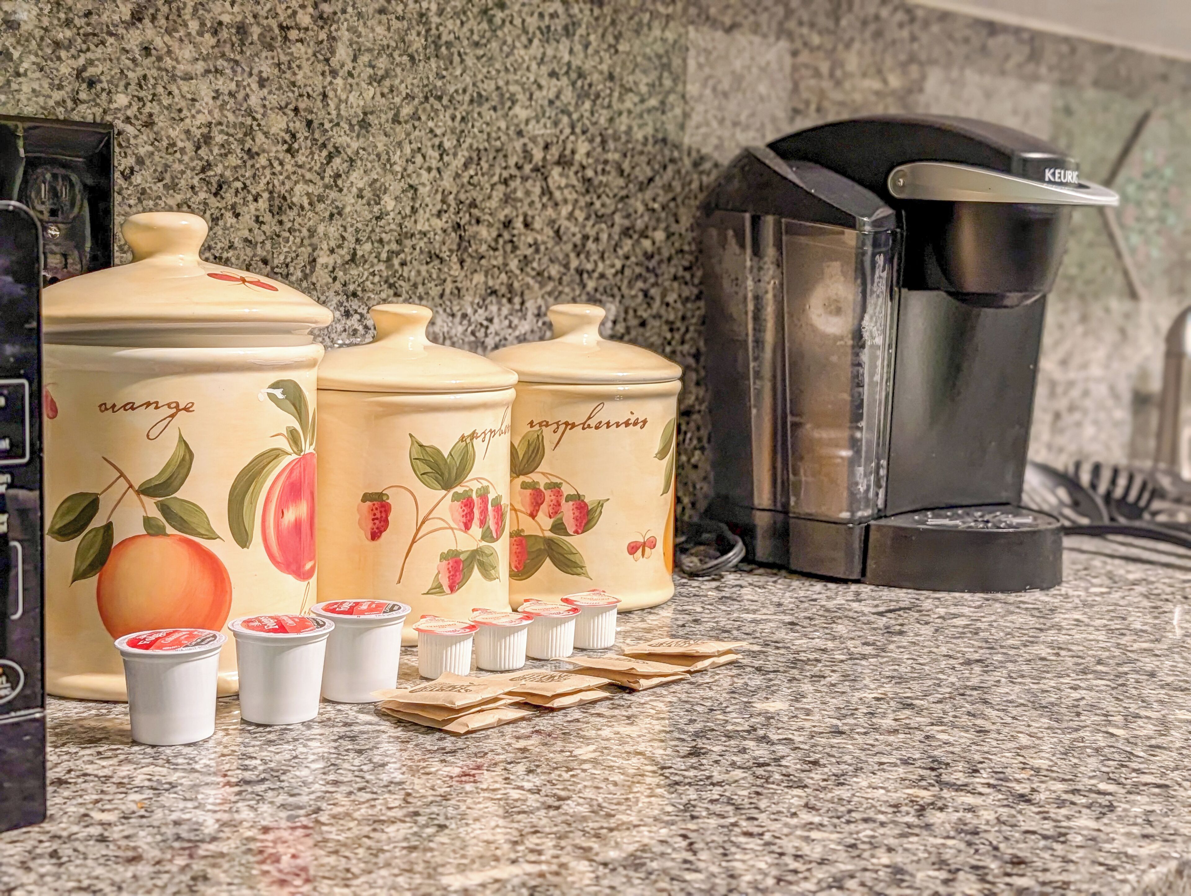 Kitchen storage containers with fruit designs, a coffee maker, and disposable cups on a granite countertop.