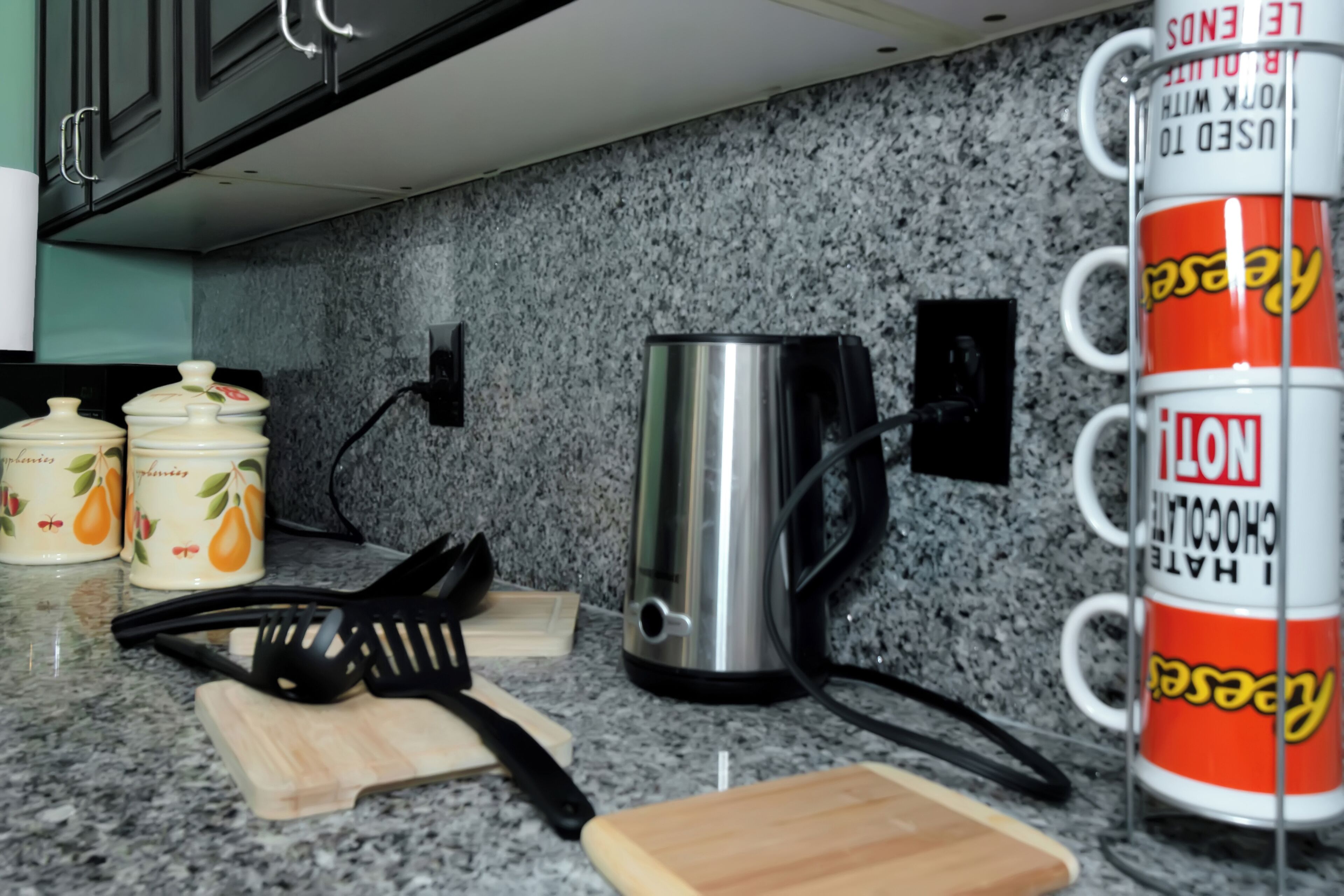 A kitchen counter with a stainless steel coffee pot, a red mug with yellow text, and a wooden cutting board.