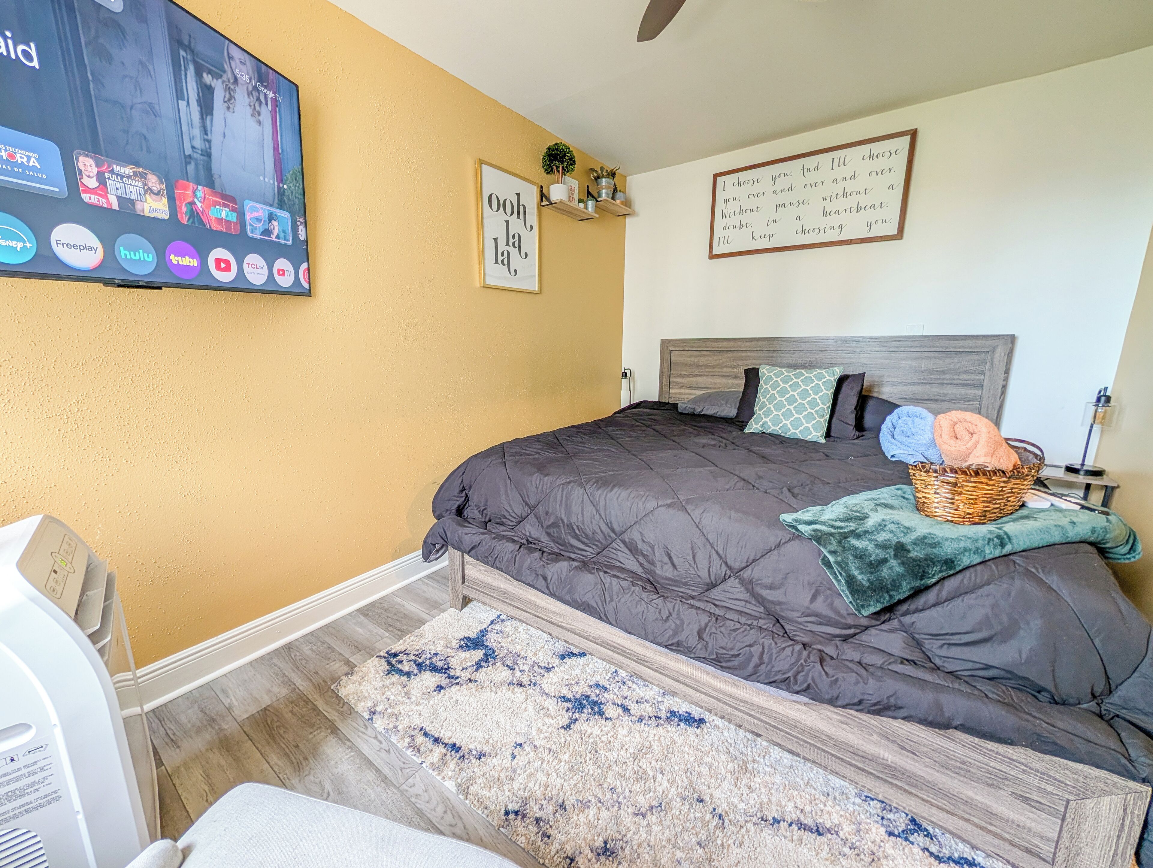 A bedroom with a bed, a TV mounted on the wall, a bedside table with a lamp, and a wall-mounted shelf with decorative items.