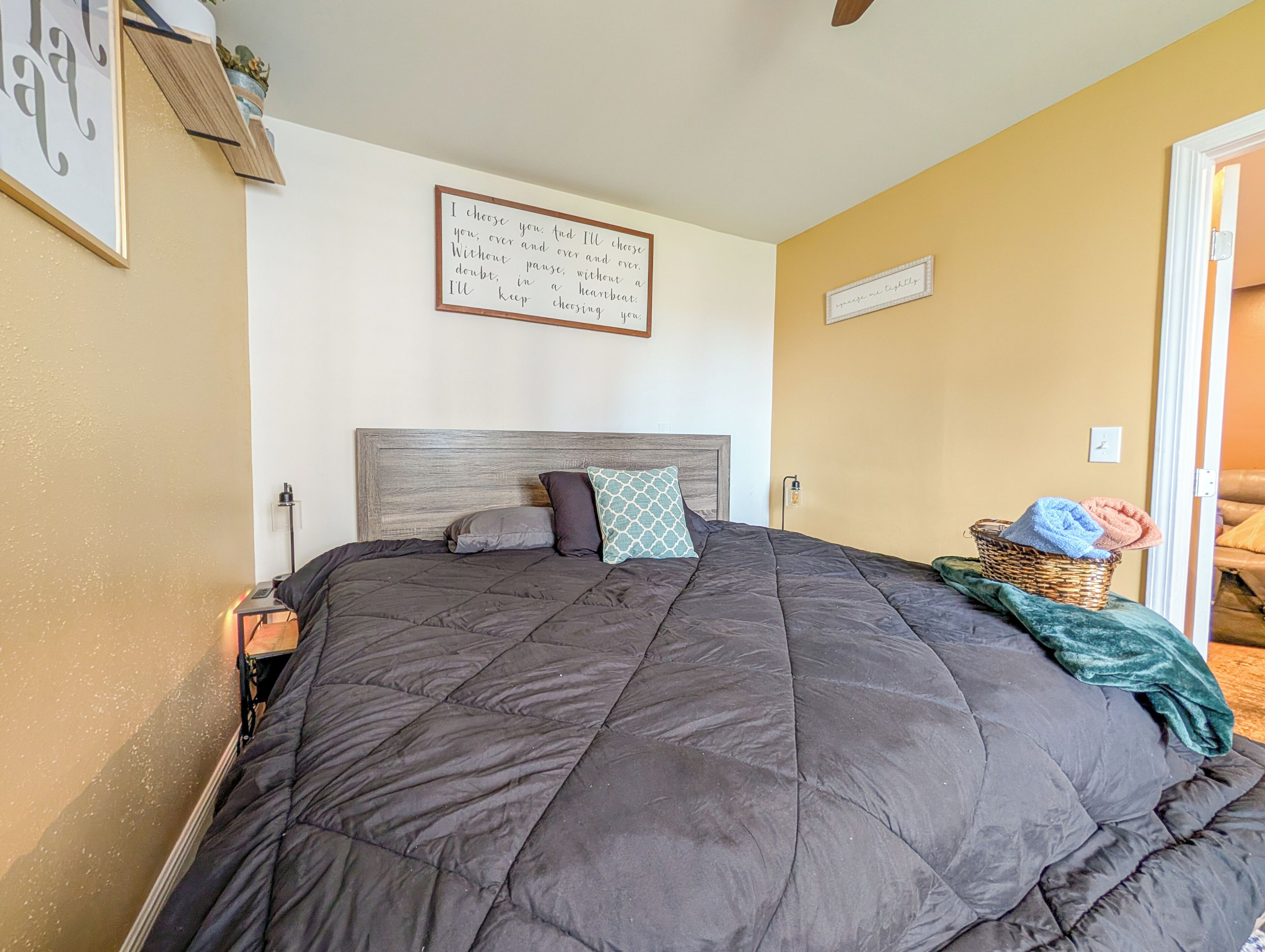A bedroom with a large bed, a nightstand, a wall-mounted light, a framed inspirational quote, and a wicker basket with towels.