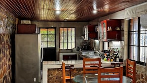 A rustic kitchen with wooden ceiling beams, a stone wall, and a dining area with a glass table and wooden chairs.