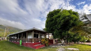 A traditional wooden building with a covered porch, surrounded by lush greenery and a mountain backdrop.