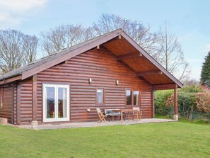 A log cabin with a large sliding glass door, a covered patio with a table and chairs, and a sloped roof. - 2 bedroom accommodation in Burlescombe (Burlescombe)
