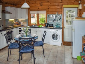 A compact kitchen with white cabinets, a dining table with blue chairs, a washing machine, and a window with a view of greenery. - 2 bedroom accommodation in Burlescombe (Burlescombe)
