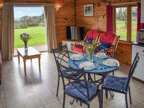 Een eetgedeelte met een ronde tafel voor vier personen, omringd door stoelen met blauwe kussens. Een televisie is aan de muur bevestigd, en een raam biedt uitzicht op het omliggende landschap.