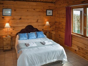 A wooden cabin bedroom with a bed, two bedside tables with lamps, a window with curtains, and two framed pictures on the wall. - 2 bedroom accommodation in Burlescombe (Burlescombe)
